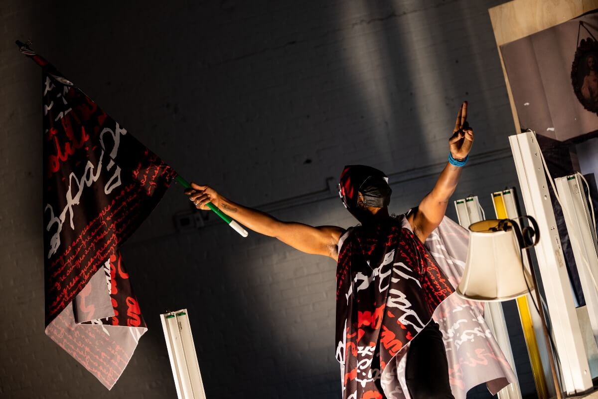 A dark-skinned audience member stands with arms up wrapped in flags that read: sangre revolucionaria, paz and victoria. Their left hand is in a peace sign. Their right hand holds up another flag on a broomstick.