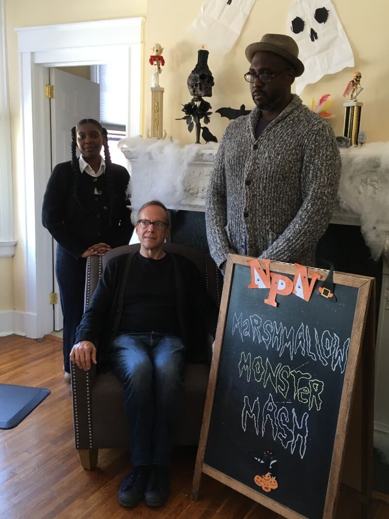 Three adults pose solemnly in front of a white fireplace mantle for a different photo than the one we are looking at; they are all looking off to the side at an unseen camera. Although they look serious, there is an air of playfulness and exaggeration to the poses. The fireplace mantle is covered in clumps of white Poly-Fil fiber meant to suggest spider webbing. On top of the mantle are other Halloween decorations, including a skull with a black candle on top, the silhouette of a raven or other black bird, the silhouette of a bat, and black roses. In the foreground next to the group is a white sandwich chalk board with cut-out, orange-and-white paper letters that spell NPN affixed to the top, next to a paper cutout of a witch's hat. On the chalkboard in squiggly hand-drawn spooky lettering are the words "Marshmallow Monster Mash." Of the three people posing, on the left is a black woman wearing a white collar shirt and long-sleeve, black, buttoned sweater and dark blue jeans. She has a medium thickness braid on each side of her head. She is standing partially behind a brown leather high-backed chair, where there sits a white man with short, lightly colored hair and black horn-rimmed, half-frame glasses. He is wearing a black shirt and an opened long-sleeve, black sweater or jacket, and blue jeans. The third figure, on the right and standing next to the chair and behind the sandwich board, is a tall black man with a closely trimmed black goatee and tortoise shell glasses. He is wearing a light brown, pork pie hat, and he has a black and white textured cardigan that's buttoned up, with what appears to be a thin, dark gray or black shirt underneath. his legs are obscured by the sandwich board.