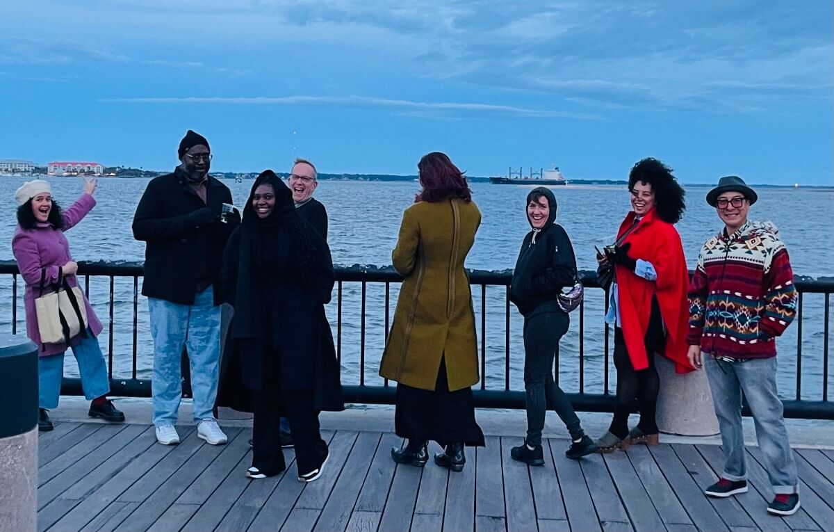 Eight adults of various skin tones stand outside on a pier or dock with a dark metal railing. Behind them, on the other side of the railing, is a harbor with a boat in the distance and a shoreline in the far distance. The day is slightly overcast but the sky is blue nearer the horizon. It appears to be winter, because everyone is wearing warm clothing, coats, scarves, and hats. The are all laughing and smiling but they are not posing together for an official photograph. It looks more like they were caught slightly off guard by the photographer and all but one of them is turned to stare into the camera for the casual photo opportunity.